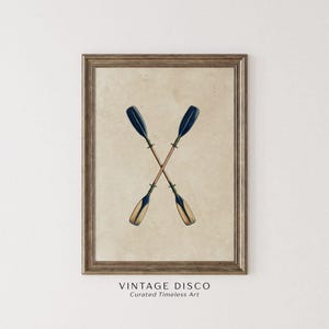 Puede incluir: Impresión enmarcada con dos remos cruzados. Los remos tienen palas azul oscuro y mangos marrón claro, sobre un fondo beige. El marco es de color marrón oscuro. El texto "VINTAGE DISCO Curated Timeless Art" está en la parte inferior.