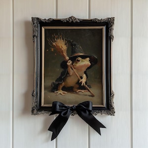 May include: A framed painting of a frog wearing a black witch hat and cape, holding a broomstick. The artwork is encased in an ornate, dark-toned frame with gold accents and features a large, black satin ribbon bow attached to the bottom center.