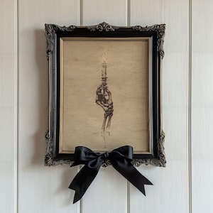 May include: A framed art print featuring a detailed ink illustration of a skeletal hand holding a lit candle. The artwork is set on a tan background within an ornate black and gold frame, accented with a large, decorative black satin ribbon bow.