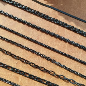 May include: A set of eight black chain necklaces of different styles and widths. The chains are laid out in a row on a brown surface.