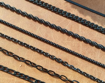 May include: A set of eight black chain necklaces in different styles and widths. The chains are laid out in a row on a brown surface.
