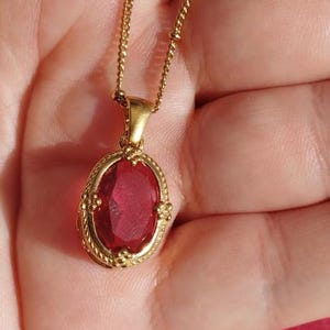 Pode incluir: Um colar de corrente de ouro com um pingente de pedra oval vermelha. O pingente está engastado em uma moldura dourada com um design detalhado.