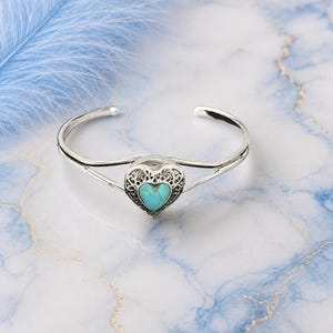 May include: A silver cuff bracelet with a heart-shaped turquoise stone. The heart is framed by an ornate, openwork design. The bracelet is displayed on a marble surface with a blue feather in the background.