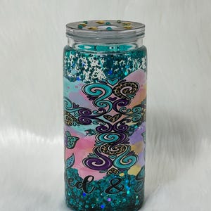 May include: A clear, cylindrical tumbler with a teal glitter base and lid. The tumbler features a colourful design with swirls, hearts, and the words "seed & feed". The design includes pink, purple, and teal watercolour-like elements.