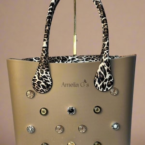 May include: A beige tote bag with a leopard print lining and handles. The bag has a variety of decorative buttons and the text "Amelia G's" on the front.