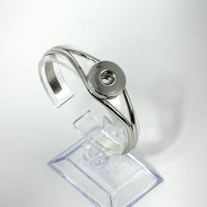 May include: A silver-toned bracelet with a large, circular snap closure. The bracelet has a sleek, modern design with a curved band. It is displayed on a clear, plastic stand against a white background. The bracelet appears to be made of metal.