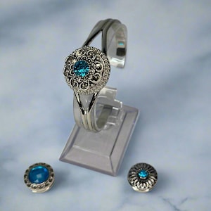May include: A silver-toned bracelet with a large, round, ornate centerpiece featuring a bright blue gemstone. Two matching snap buttons with blue gems are also present. The bracelet is displayed on a clear stand.