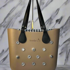 May include: A beige tote bag with black leather handles and a black and white houndstooth lining. The bag has a silver logo that reads "Amelia G's" and is adorned with silver, sparkly, decorative studs.