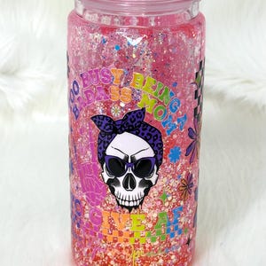 May include: A pink glitter tumbler with a screw-on lid. The tumbler features a skull graphic wearing sunglasses and a bandana, with the text "Too Busy Being a Badass Mom" and "Give 'Em Hell".