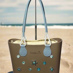 May include: A brown tote bag with blue handles and a white lining. The bag is decorated with various blue and turquoise embellishments, including a starfish, a turtle, and seashells. The bag has the text "Amelia G's" printed on the front.