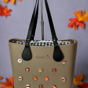 May include: A taupe tote bag with black handles and decorative pins. The bag features a black and white houndstooth lining. The pins have fall-themed designs, including pumpkins and plaid patterns. The text "Amelia G's" is visible on the bag.