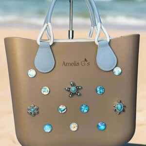 May include: A beige tote bag with light blue handles and a white rope detail. The bag has a variety of blue and turquoise embellishments, including a starfish, a turtle, and a flower. The bag is labeled "Amelia G's".