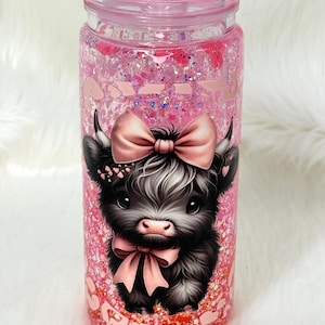May include: A pink glitter tumbler with a cartoon Highland cow wearing a pink bow. The tumbler has a pink lid and is filled with pink glitter and small decorations. The cow has black and white fur and a pink nose.