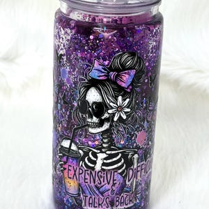 May include: A purple glitter tumbler with a decorative lid. The tumbler features a skeleton graphic wearing sunglasses and holding a drink, with the text "Expensive & Difficult Talks Back". The lid is adorned with daisy and flower accents.
