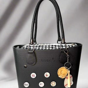 May include: Black tote bag with a black and white houndstooth lining. The bag has a black leather handle and a silver charm with a baseball and bat. The bag is decorated with various colorful buttons.