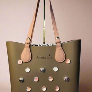 May include: A brown tote bag with pink straps and a white logo that says "Amelia G's". The bag is decorated with small, colorful buttons.
