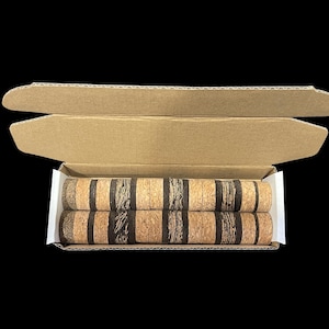May include: Two cylindrical cork products with dark brown stripes are displayed inside a cardboard box. The box is open, revealing the corks, which have a natural, light brown color. The background is solid black.
