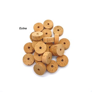 May include: A collection of natural cork beads with a central hole, scattered on a white background. The beads are cylindrical and circular, with a light brown color and visible texture. The word "Extra" is printed in black.