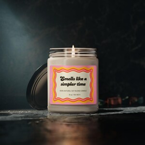 May include: A lit soy blend candle in a clear glass jar with a black lid. The label reads "Smells like a simpler time" in a retro font, framed by a pink and yellow border. The candle is 9 oz and burns for 50-60 hours.