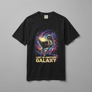 May include: Black t-shirt featuring a colorful graphic of an astronaut reading a book in space. The design includes vibrant purples, blues, and yellows, with the text "LOST IN ANOTHER GALAXY" in gold.