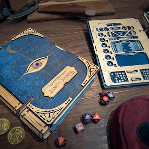 Puede incluir: Un diario de madera con una cubierta azul con un diseño celestial, un ojo y el texto "Janet's Journal". Junto a él, un tablero de juego de madera con varias ranuras y marcas. Monedas de oro y dados de múltiples caras están dispersos.