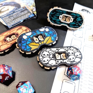 Op de afbeelding: Houten spel tellers met roterende nummer displays. Ontwerpen omvatten houtnerf, blauw mozaïek, groene stof en een schedelpatroon. Dobbelstenen en een spelkaart zijn ook aanwezig, wat wijst op een tabletop game thema.