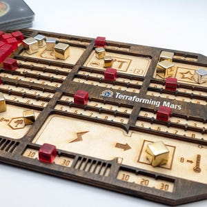 Puede incluir: Un componente de juego de mesa de madera con el texto "Terraforming Mars". El tablero presenta líneas grabadas, números y símbolos, con piezas de juego rojas y doradas. Una baraja de cartas es visible en el fondo.