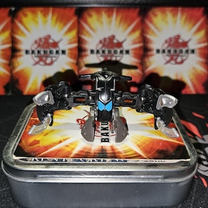 Peut inclure: Un jouet Bakugan noir et argent avec des yeux bleus. Le jouet est dans une boîte en métal avec un logo Bakugan et un fond rouge et orange.