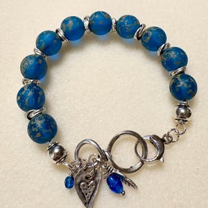 May include: A blue beaded bracelet with silver accents. The bracelet features large, round blue beads with a mottled gold pattern. Silver rings and beads separate the larger beads. The bracelet has a silver clasp and charms, including a heart and a feather.