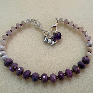 May include: A beaded bracelet featuring faceted light pink and deep purple stones separated by small dark beads. The bracelet includes a silver-toned lobster claw clasp, a leaf-shaped charm, a small butterfly charm, and a single dark purple bead.
