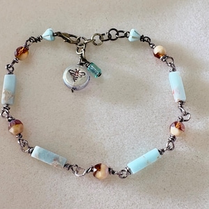 May include: A beaded bracelet featuring light blue and brown beads, connected by a dark metal chain. A small charm with a bee design and a small blue bead dangle from the bracelet. The bracelet is designed for wrist wear.