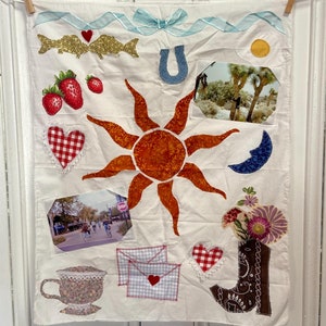 May include: A white fabric square with various appliques, including a red and orange sun, a horseshoe, a red and white checkered heart, a blue crescent moon, a brown boot, two red strawberries, two gold fish, a blue ribbon, a white envelope with a red heart, and two photographs.