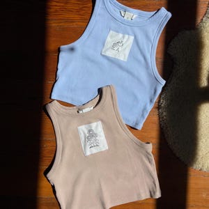 以下が含まれることがあります： 木製の表面に、ライトブルーとタンの2色のリブ編みタンクトップが2枚並んでいます。各トップスには、白地に白黒の花柄が施された正方形のパッチが付いています。トップスはノースリーブで、丸いネックラインです。