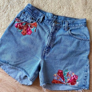 May include: Light blue denim shorts with raw hems. The shorts feature colorful floral fabric patches on the front pockets and the lower right side. The shorts have a button closure and classic five-pocket design.