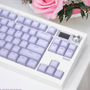May include: A white mechanical keyboard with lavender keycaps. The keyboard features a number pad, function keys, and a row of media keys. The keyboard is on a white surface.