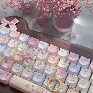 Może przedstawiać: Niestandardowa klawiatura z nakładkami na klawisze z motywem niedźwiedzia w pastelowych kolorach. Nakładki na klawisze przedstawiają różne wzory niedźwiedzi i są ułożone na drewnianej podstawie. Obecna jest również różowa kokarda i wazon z różowymi kwiatami.