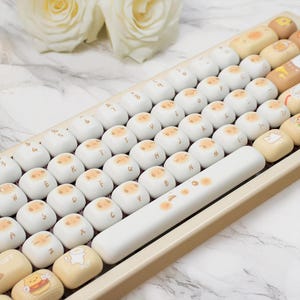 May include: A beige and white mechanical keyboard with custom keycaps. The keycaps are white with a light orange design. The space bar is white with orange accents. Some keycaps have cartoon illustrations. The keyboard is angled.