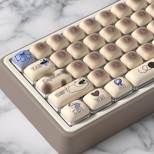 Könnte beinhalten: Nahaufnahme einer mechanischen Tastatur mit beigefarbenen Tastenkappen und einem grauen Rahmen. Die Tastenkappen zeigen verschiedene Symbole und Text, darunter eine Katze, ein Herz mit "WIN" und das Wort "TAB". Die Tastatur liegt auf einer marmorierten Oberfläche.