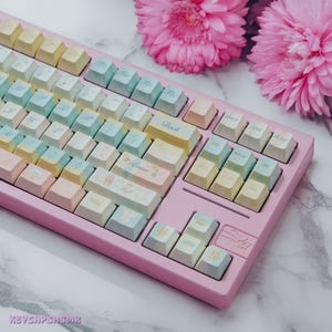 Może przedstawiać: Klawiatura mechaniczna w pastelowych kolorach z różową podstawą i nakładkami w odcieniach żółtego, niebieskiego i różowego. Klawiatura posiada niestandardowe nakładki z motywami kwiatowymi i gwiazdami. Słowo "Back" jest widoczne na jednej z nakładek. Dwa różowe kwiaty w tle.