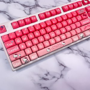 May include: A pink and white keyboard with Hello Kitty keycaps. The keyboard has a white base and pink keys with white lettering. The keys are arranged in a standard QWERTY layout. The keyboard has a Hello Kitty logo on the space bar and a Hello Kitty character on the Enter key.