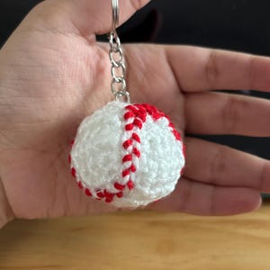 May include: A handmade baseball keychain. The baseball is white with red stitching, crafted from yarn. It is attached to a silver-colored chain and keyring. The keychain is a sports-themed accessory.