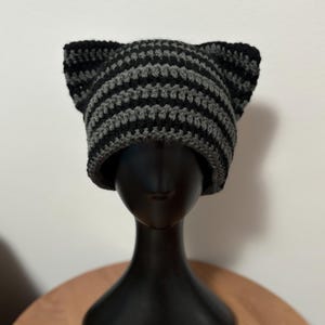 May include: A handmade crochet hat with cat ears. The hat features alternating horizontal stripes of black and gray yarn. The hat is displayed on a black mannequin head.