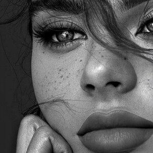 May include: Black and white close-up portrait of a person's face. The image highlights the eye, nose, and lips. The person has long eyelashes, full lips, and freckles. Hair partially obscures the face, and a hand rests on the chin.