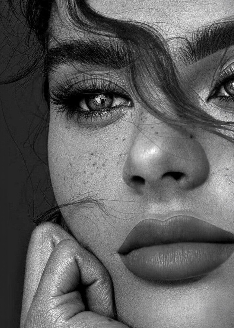 May include: Black and white close-up portrait of a person's face. The image highlights the eye, nose, and lips. The person has long eyelashes, full lips, and freckles. Hair partially obscures the face, and a hand rests on the chin.