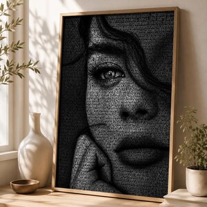 May include: Black and white portrait art print of a woman's face, with the image composed of small text. The artwork is framed in a light brown wooden frame. The woman's eye is in focus, and her hand is touching her face.