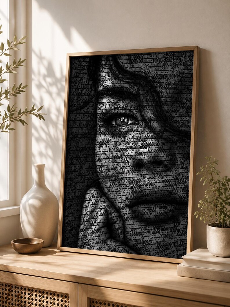 May include: Black and white portrait art print of a woman's face, with the image composed of small text. The artwork is framed in a light brown wooden frame. The woman's eye is in focus, and her hand is touching her face.