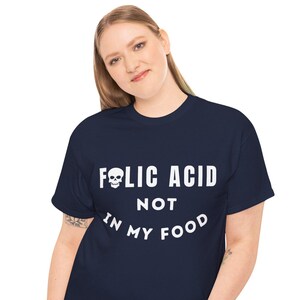 May include: Navy blue t-shirt with the text "FOLIC ACID NOT IN MY FOOD" in white, featuring a skull graphic. The shirt is worn by a person in blue jeans.