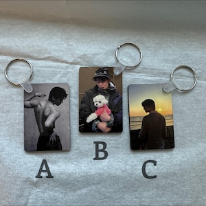 May include: Three keychains with different images. The first keychain features a black and white photo of a person with their back to the camera. The second keychain features a color photo of a person holding a small white dog. The third keychain features a color photo of a person standing on a beach at sunset.