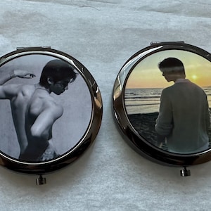 May include: Two round, dark metal compact mirrors. One mirror features a black and white image of a person with their back to the viewer. The other mirror shows a person on a beach at sunset.