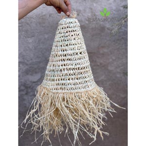 May include: A woven, cone-shaped lampshade made of natural straw. The lampshade has a fringed bottom and is held by a hand. The text "ILMASHOPM" is visible in the upper right corner.
